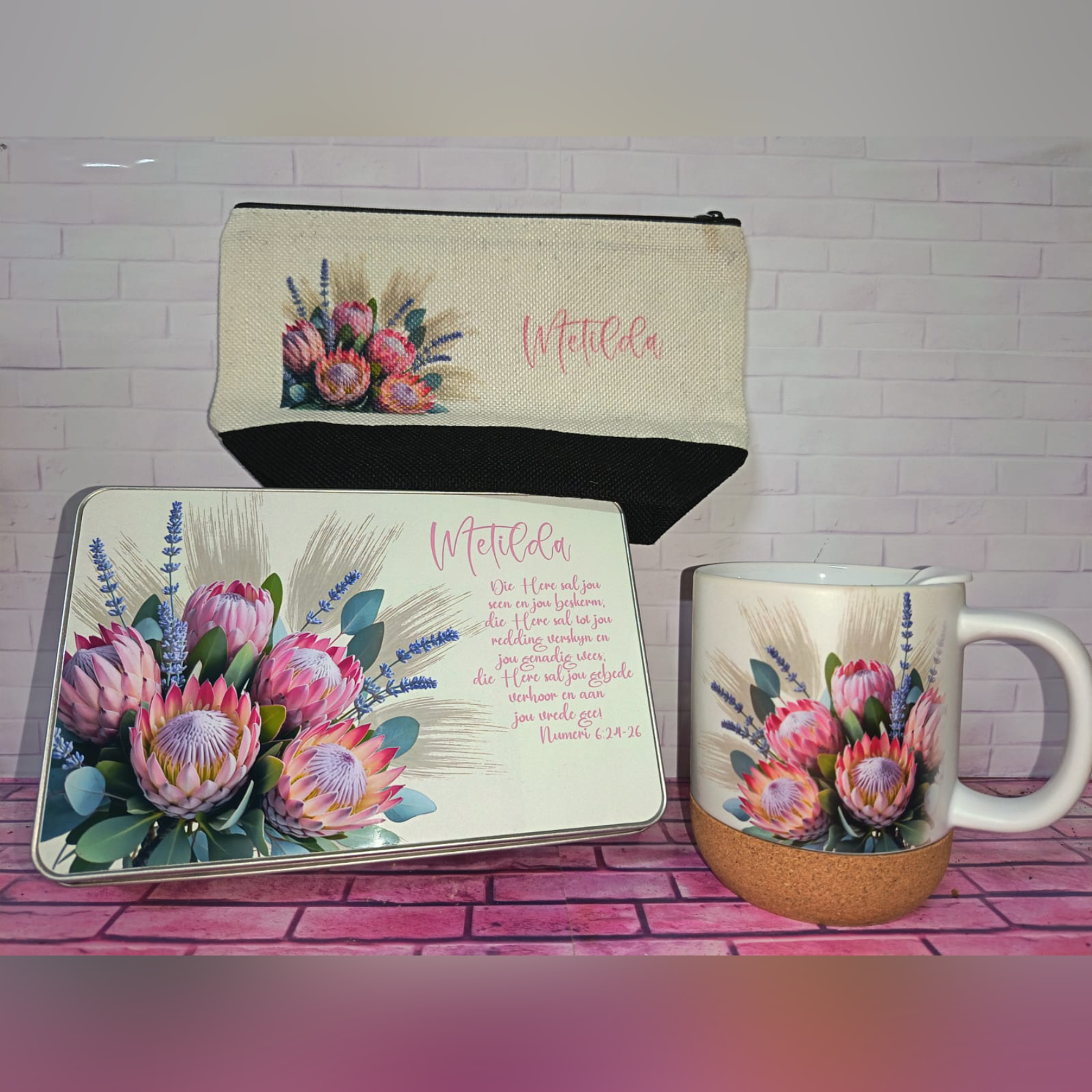 Floral-themed desk set: a metal tin, a notebook, and a mug, all with bright pink protea-style flowers and lavender on a pink brick background.