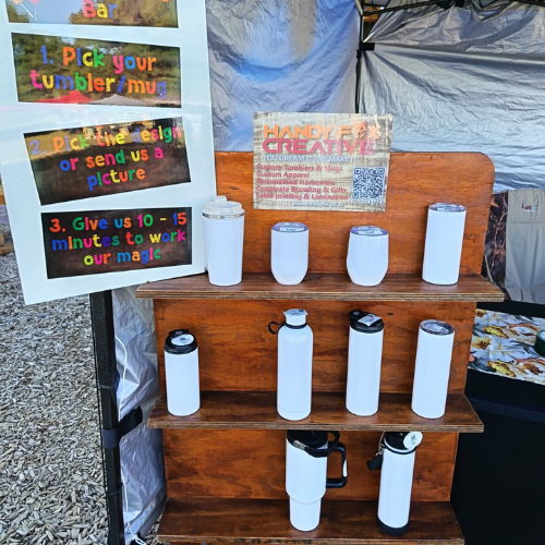 Wooden display stand inside a tent with white insulated tumblers on three shelves and colorful signs instructing customers to pick a tumbler and design the mug.