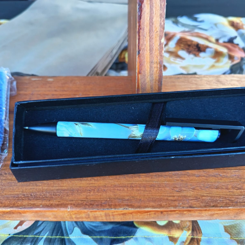 Blue decorative ballpoint pen in a black presentation box secured with a strap, resting on a wooden table.