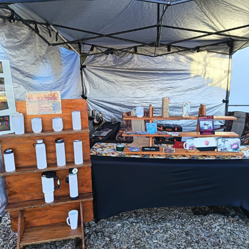 Outdoor market stall under a blue canopy; wooden shelves display white water bottles and mugs, with a table of handmade crafts nearby.