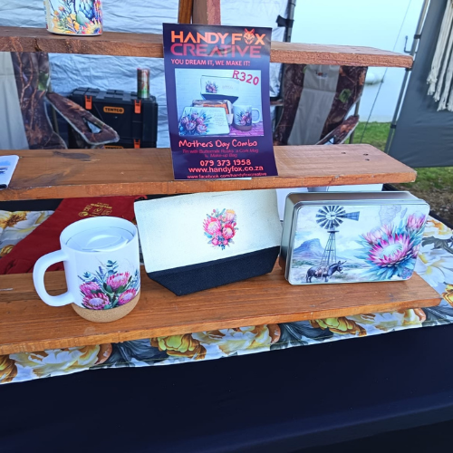 Outdoor craft stall display on wooden shelves featuring a white mug with pink floral pattern, a small white-and-navy box with a pink flower design, and a metal tin with a windmill and pink flower motif, plus a floral-printed item beneath; a sign for Handy Creative is visible in the background.