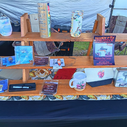 Wooden market stall shelf displaying painted jars, a floral mug, greeting cards, and a Handy Creative flyer.